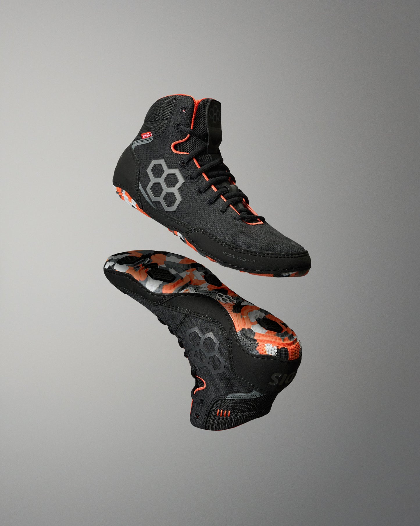 A pair of athletic high-top wrestling shoes floating against a neutral gray gradient background. The shoes are primarily black with vibrant orange accents and a gray geometric hexagonal logo on the side.