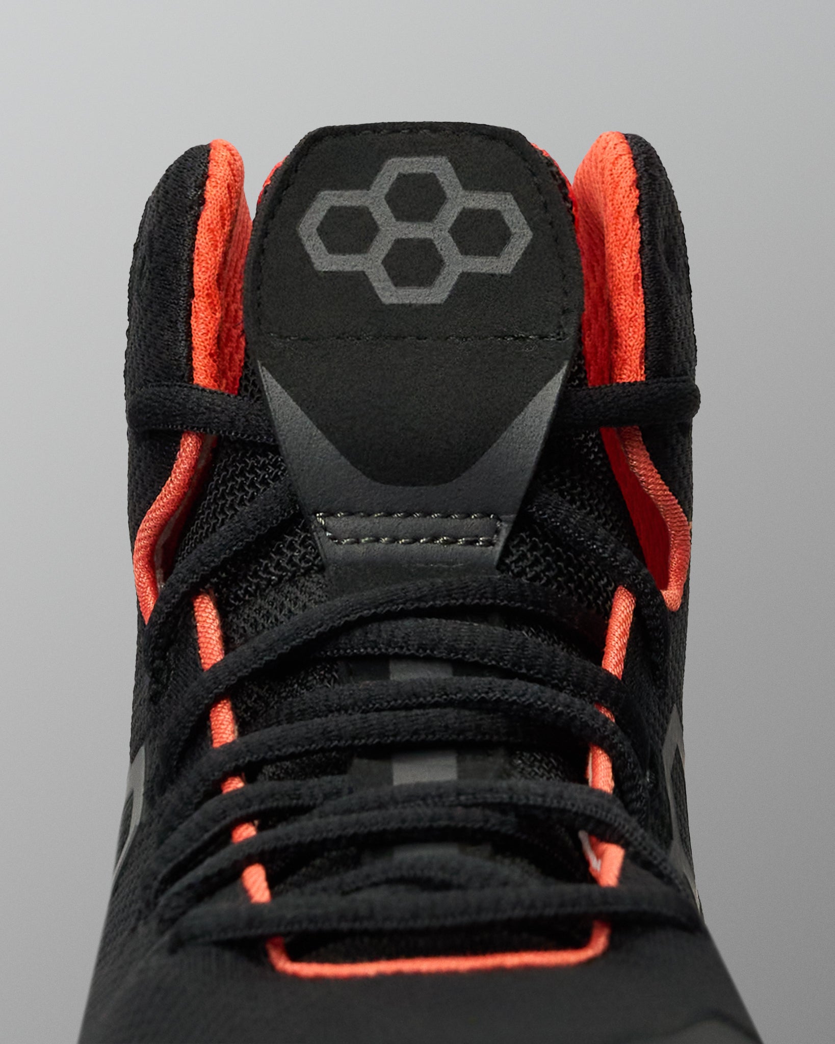 Close-up of a black high-top Wrestling Shoe with bright orange lining and laces, featuring a gray geometric logo on the tongue, set against a solid gray background.