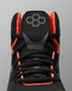 Close-up of a black high-top Wrestling Shoe with bright orange lining and laces, featuring a gray geometric logo on the tongue, set against a solid gray background.