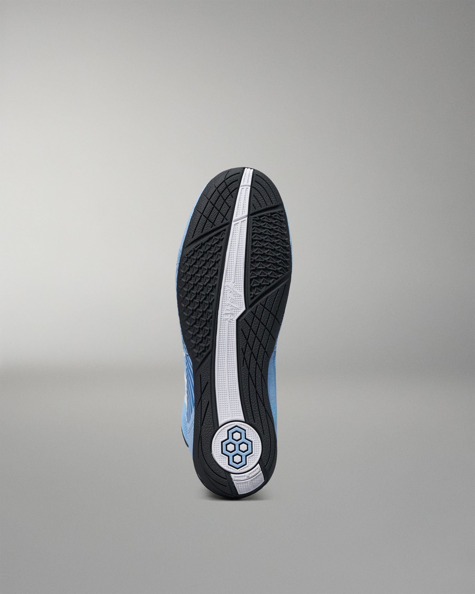 The outsole of a wrestling shoe shown from the bottom, featuring a black textured tread with a central white stripe and a circular blue and white logo near the heel, set against a neutral gray background.