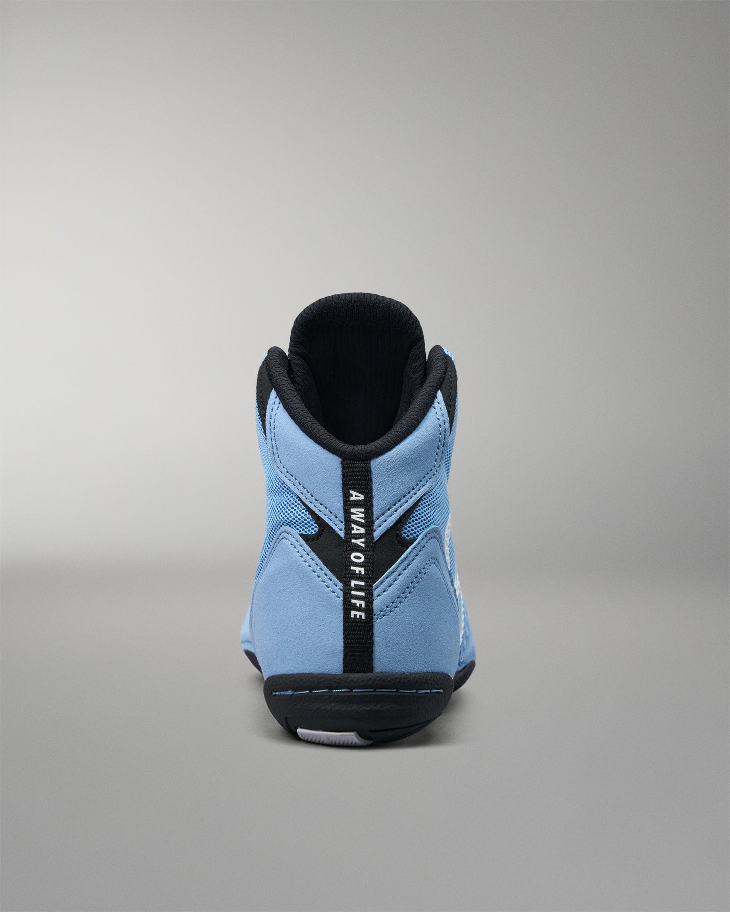A light blue high-top wrestling shoe shown from the back, featuring a black vertical strap with the text 'A WAY OF LIFE' in white, set against a neutral gray background.
