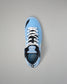 A light blue low-top wrestling shoe with black and white accents, featuring white laces and a visible RUDIS logo on the tongue and heel, photographed from a top-down angle on a neutral gray background.