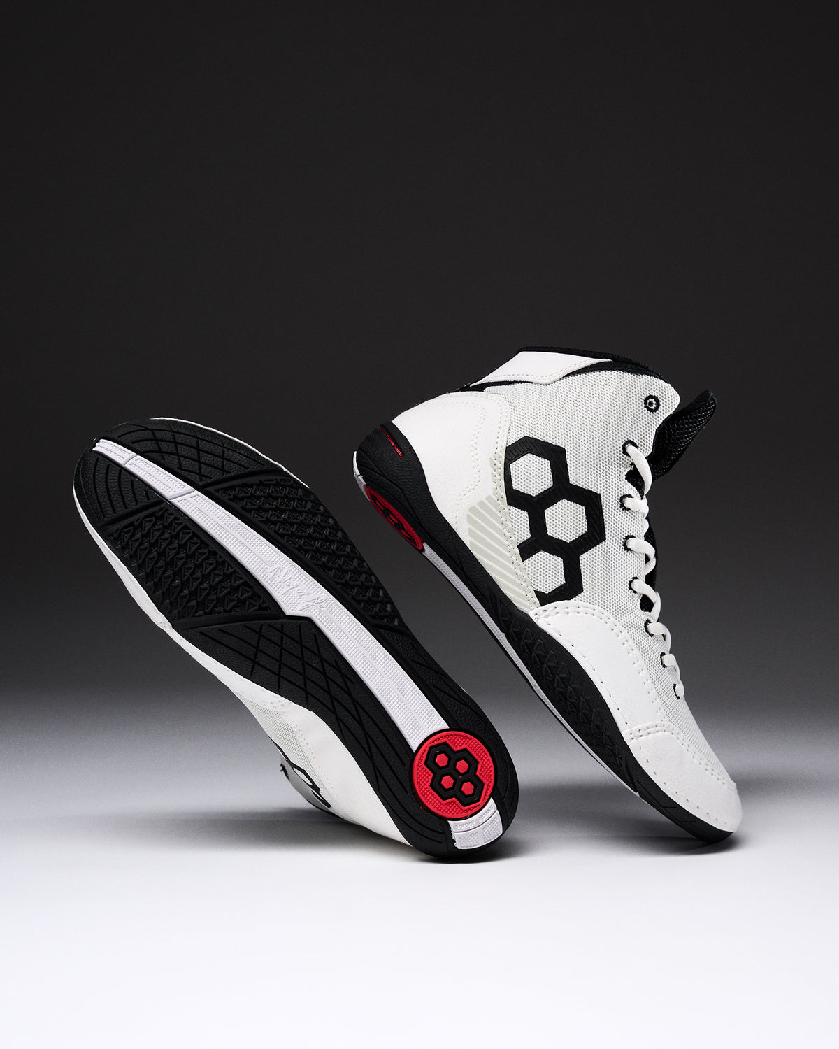 A pair of high-top wrestling shown on a dark gray to black gradient studio background. One shoe is upright showing a white leather upper with a large black geometric side logo, white laces, black trim, and a small red circular detail near the heel.