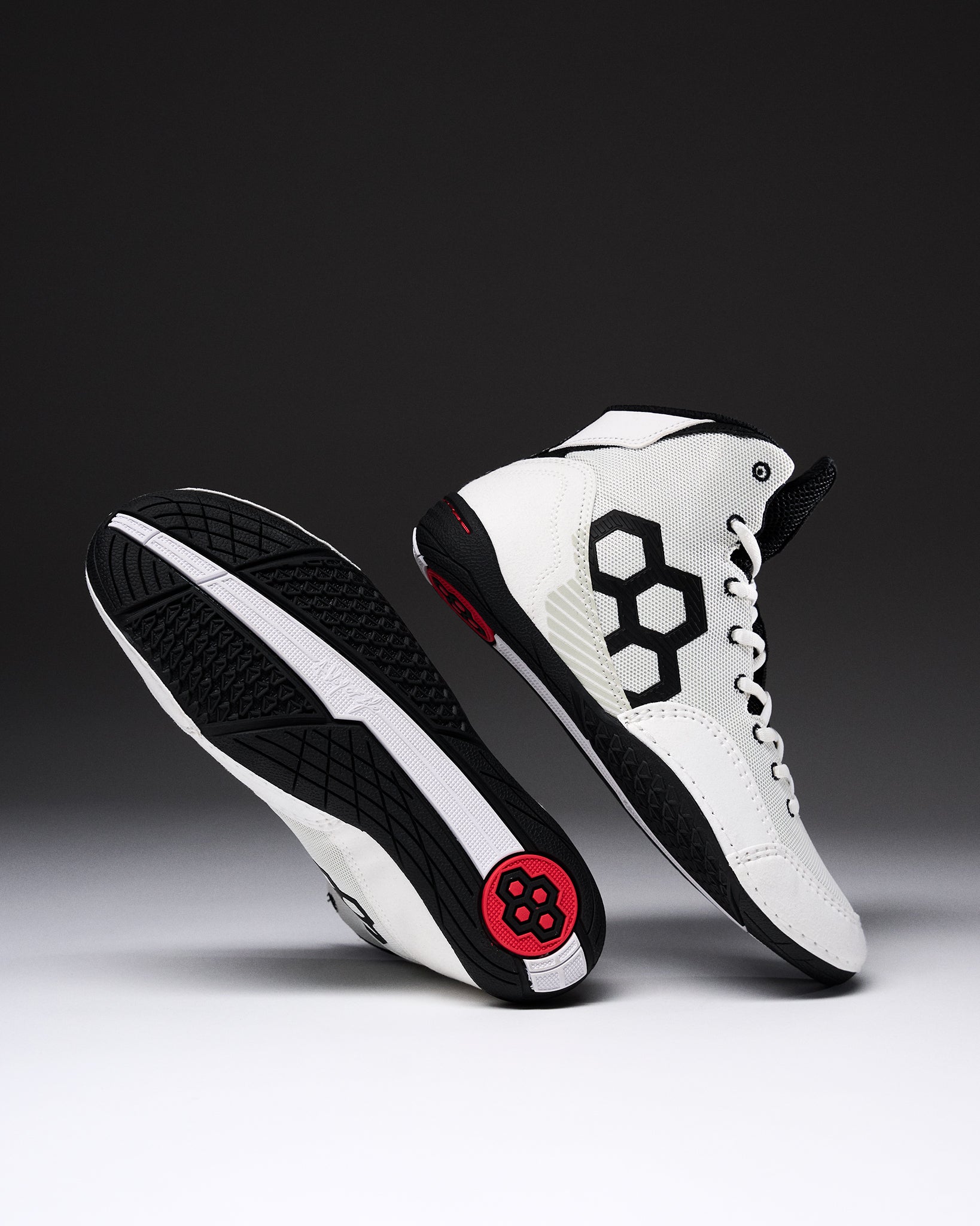 A pair of high-top wrestling shown on a dark gray to black gradient studio background. One shoe is upright showing a white leather upper with a large black geometric side logo, white laces, black trim, and a small red circular detail near the heel.