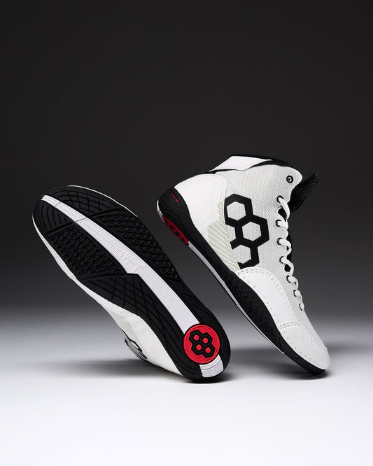 A pair of high-top wrestling shown on a dark gray to black gradient studio background. One shoe is upright showing a white leather upper with a large black geometric side logo, white laces, black trim, and a small red circular detail near the heel.