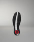 The outsole of a black wrestling shoe is shown vertically against a neutral gray background, featuring a textured black tread pattern, a central white stripe, and a red circular logo near the heel.
