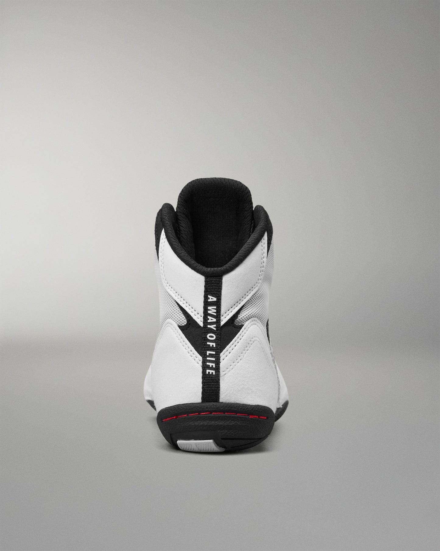 A white high-top wrestling shoe shown from the back, featuring a prominent black vertical strap with the text 'A WAY OF LIFE' in white, a black outsole, and a thin red accent line near the heel.