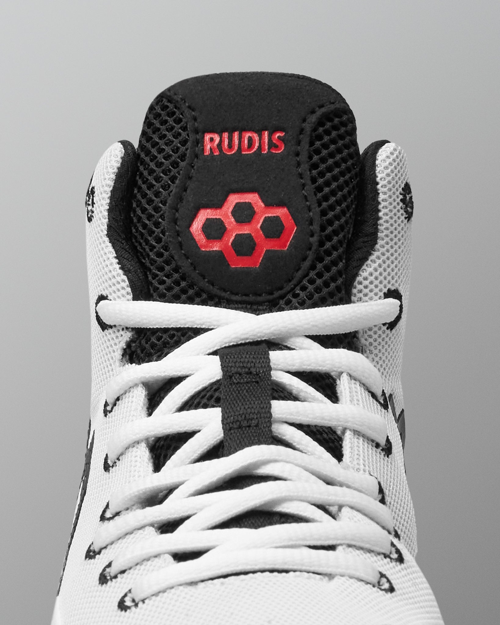 Close-up of a high-top wrestling shoe showing the tongue area, with a white mesh upper, black collar and tongue, white laces, and red embroidered branding that reads RUDIS with a red hexagon logo.