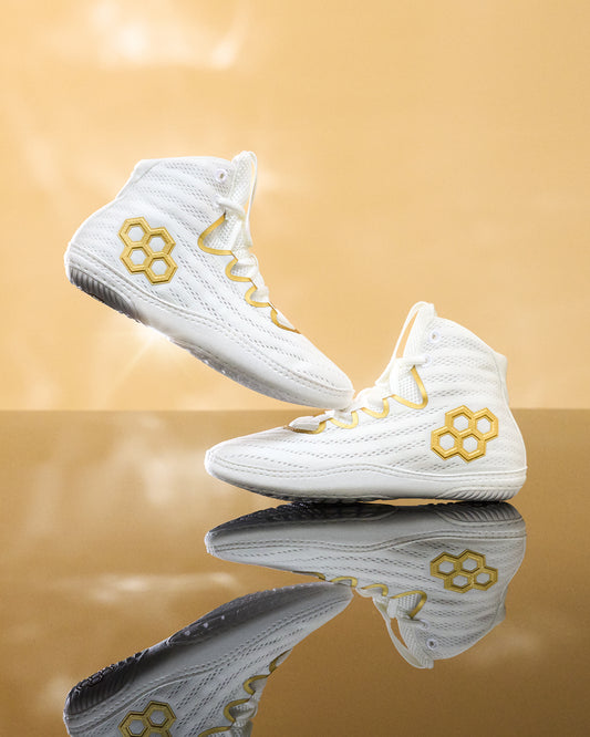 A pair of white high-top athletic wrestling shoes with gold honeycomb and wavy geometric accents on the sides, displayed on a reflective gold surface against a warm gold background.
