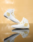 A pair of white high-top athletic wrestling shoes with gold honeycomb and wavy geometric accents on the sides, displayed on a reflective gold surface against a warm gold background.
