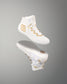 A white high-top athletic wrestling shoes with gold geometric and wavy accents, shown floating above its matching sole on a smooth gray gradient background.