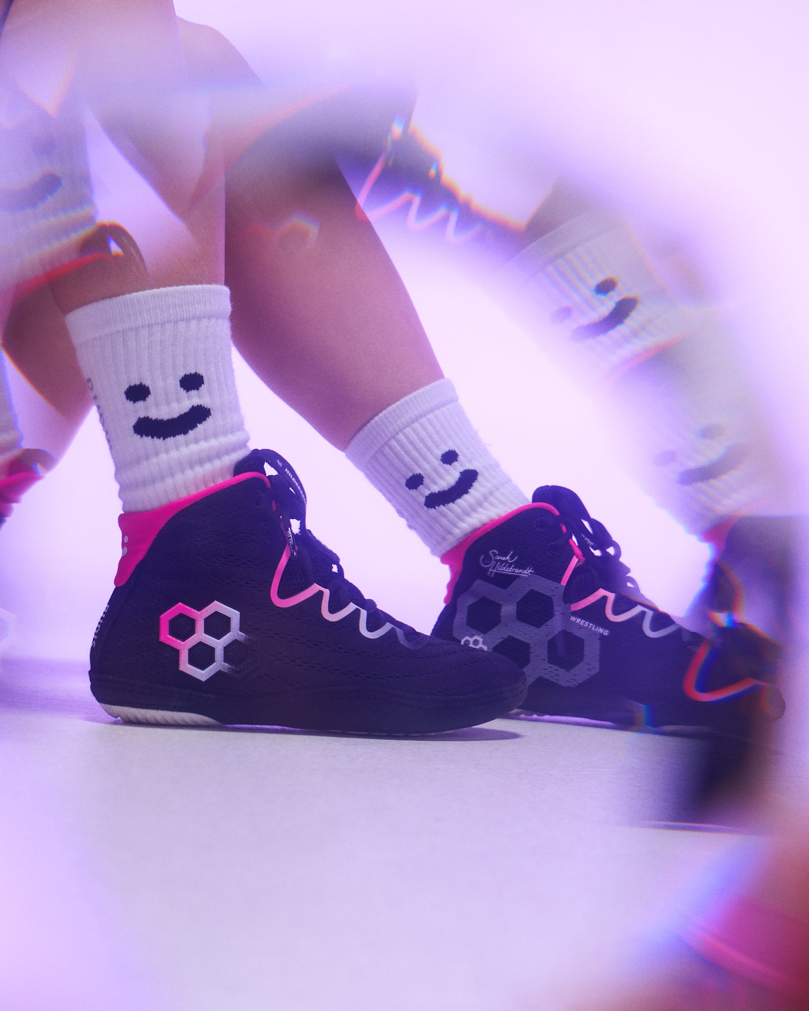 Black high-top Wrestling shoes with pink and white graphic details, worn with white crew socks featuring a smiley face, shown in a stylized purple-lit setting.