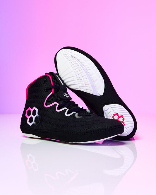 A pair of black high-top athletic wrestling shoes with vibrant pink and white accents, featuring a wavy pink stripe on the side and a white geometric logo, shown against a pink gradient background.