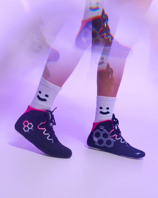 A person wearing dark navy high-top wrestling shoes with pink and gray graphic details, paired with white crew socks featuring a black smiley face, shown against a purple-lit studio background.