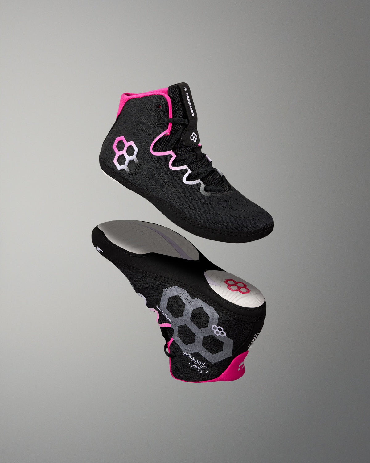 A pair of high-top wrestling shoes floating against a neutral gray gradient background. The shoes are primarily black with vibrant pink accents on the collar and sole, and gray and white geometric hexagonal patterns on the side and heel.
