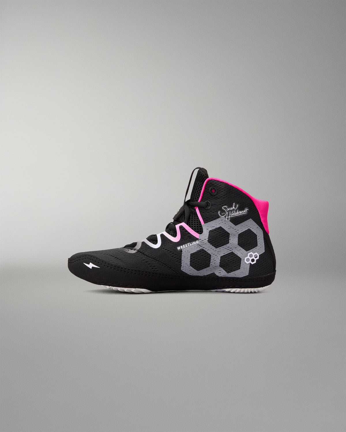 A high-top wrestling shoe in black with a bright pink collar lining, white and gray geometric side patterns, and white laces, shown against a neutral gray background.