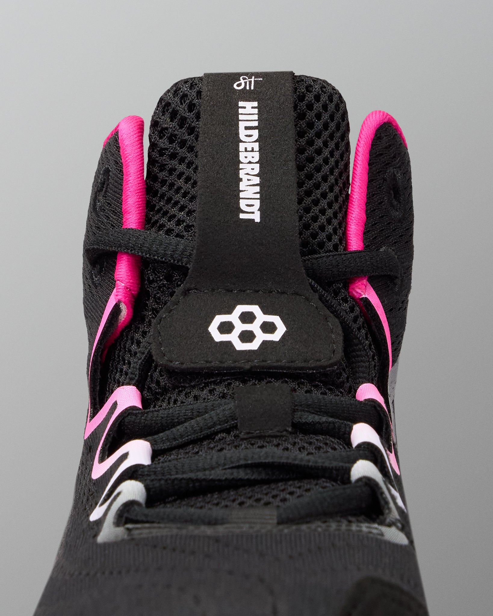 Close-up of the top of a black wrestling shoe with bright pink accents and white branding details, including the text 'HILDEBRANDT' on the tongue pull tab.