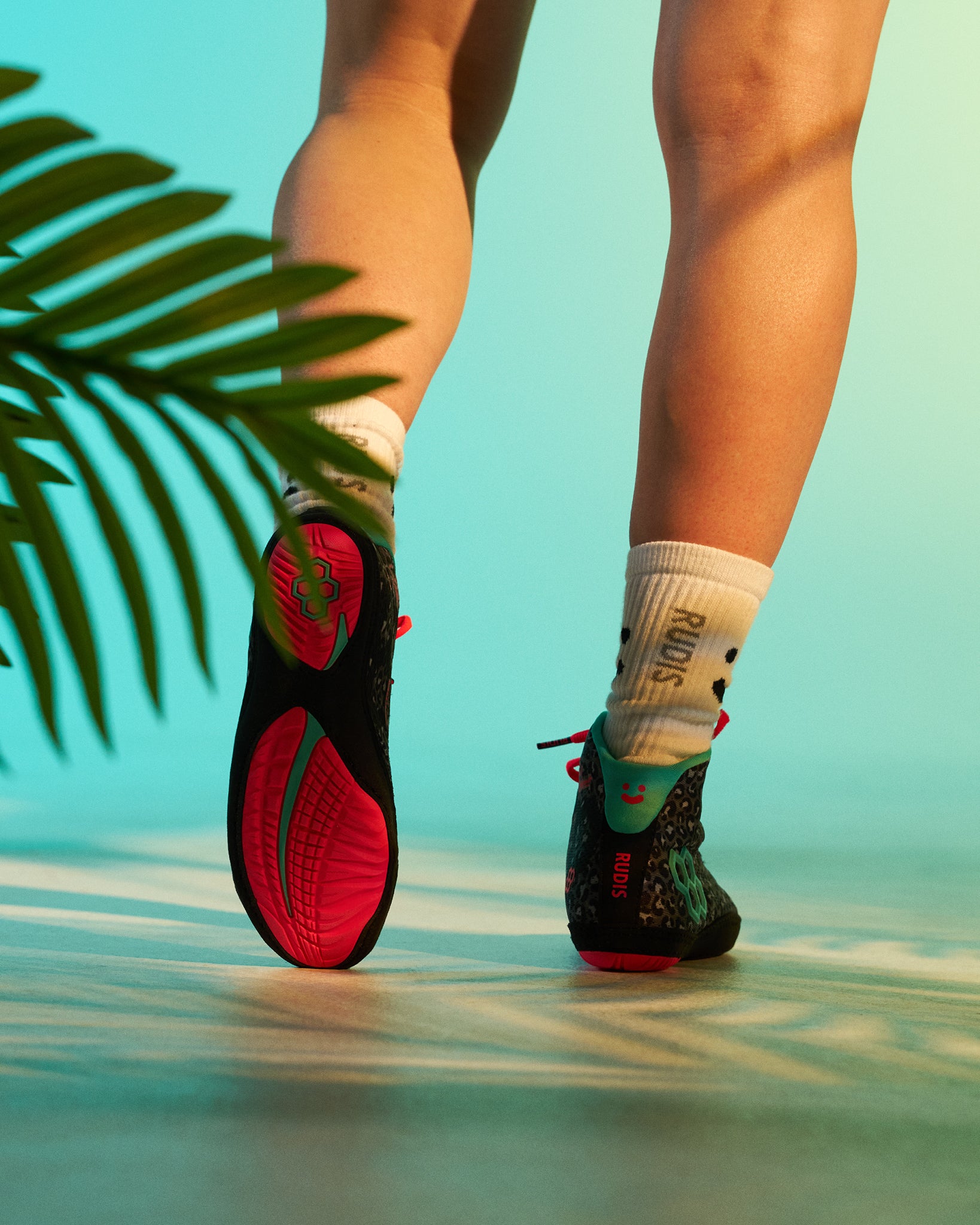Rear view of the RUDIS Hildebrandt 2.0 Girls Wrestling Shoes in Black Leopard, showcasing the bright pink outsole, teal heel accents, leopard-print upper, and RUDIS crew socks in a tropical-inspired studio setting