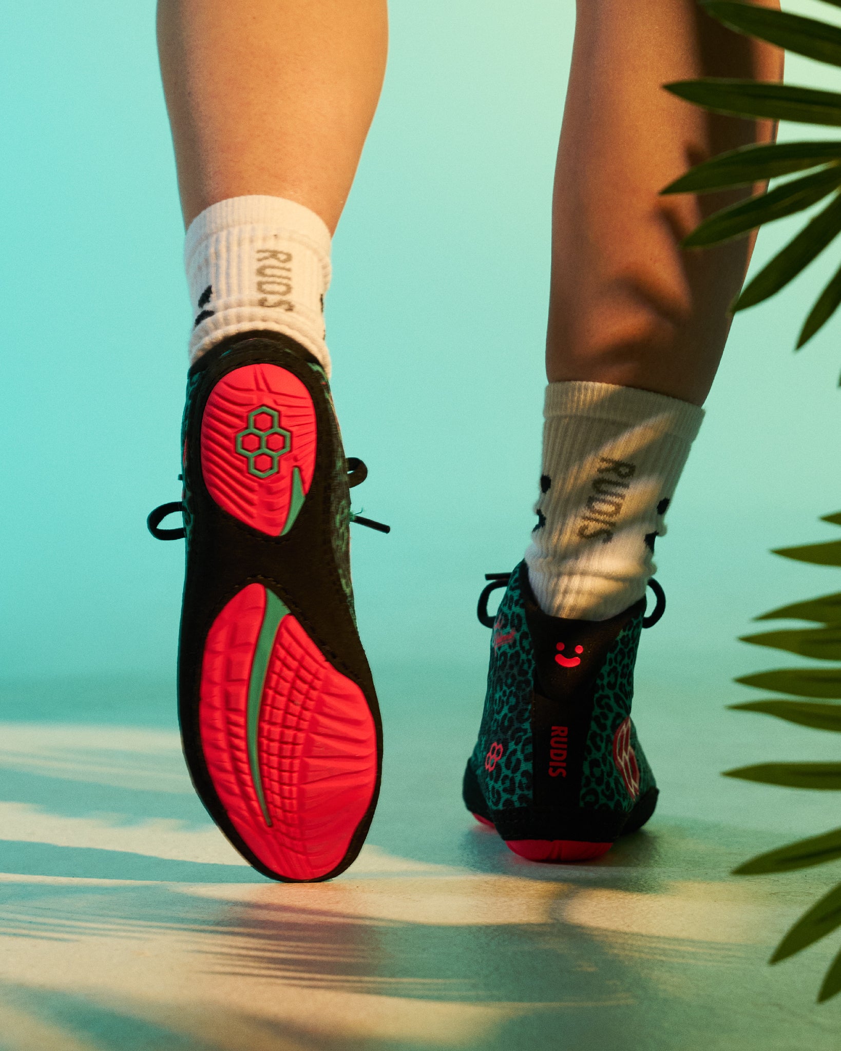 Rear view of the RUDIS Hildebrandt 2.0 Girls Wrestling Shoes in Green Leopard, highlighting the bright pink outsole, leopard-print heel, and RUDIS crew socks in a tropical-inspired studio setting.