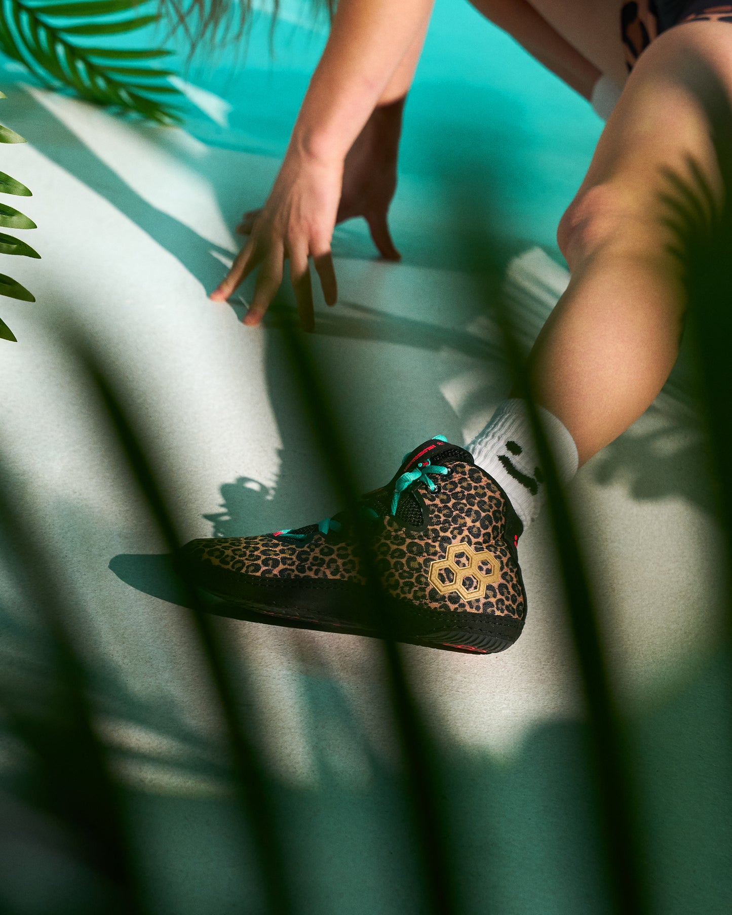 A person sitting by a turquoise pool wears a leopard-print high-top Wrestling shoe with bright teal laces and white socks, framed by palm leaves in the foreground.