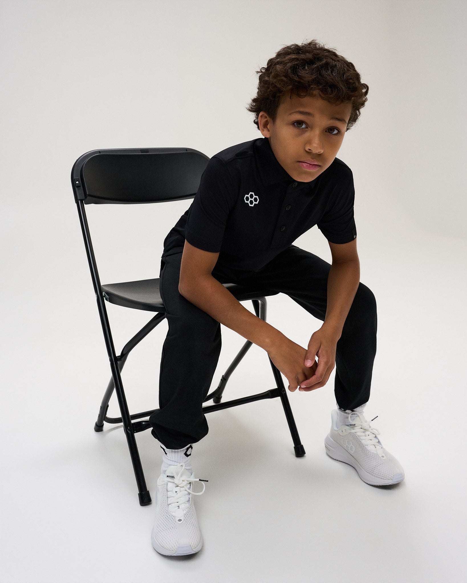 Youth athlete models the black RUDIS Textured Youth Classic Hex Polo with black athletic pants and white shoes while seated on a chair.