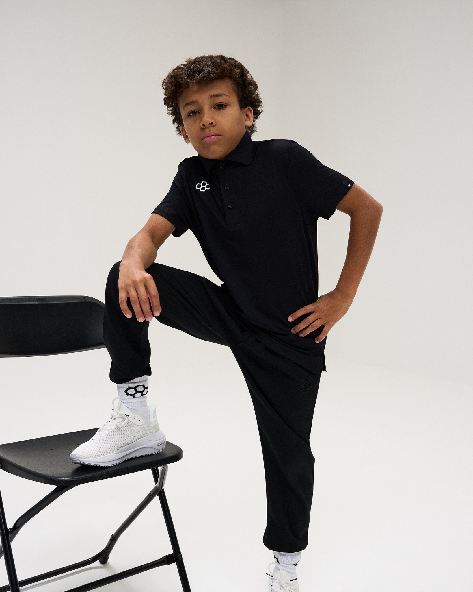 Youth athlete models the black RUDIS Textured Youth Classic Hex Polo and black athletic pants, posing with one foot on a chair.