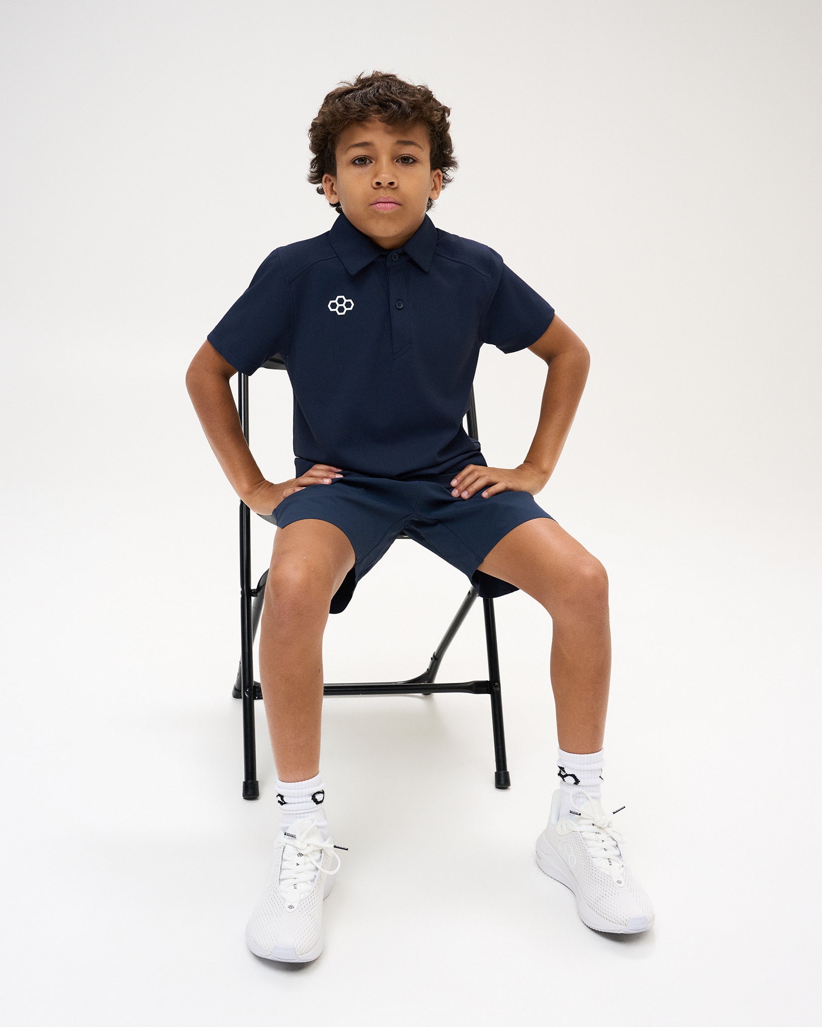 Youth athlete models the navy RUDIS Textured Youth Classic Hex Polo with matching navy shorts while seated against a white studio background.