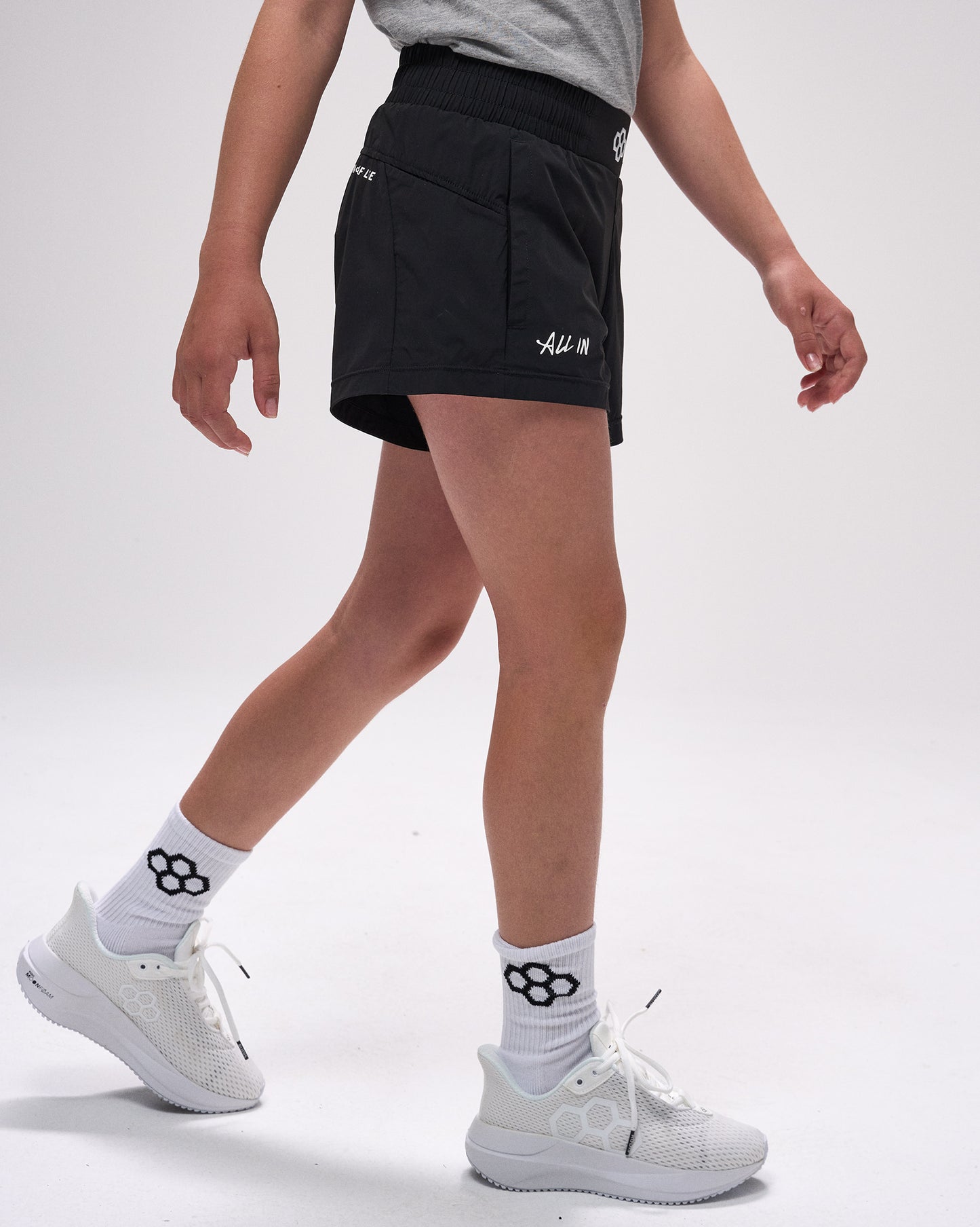Person wearing black shorts with a logo, white sneakers, and socks on a light gray background