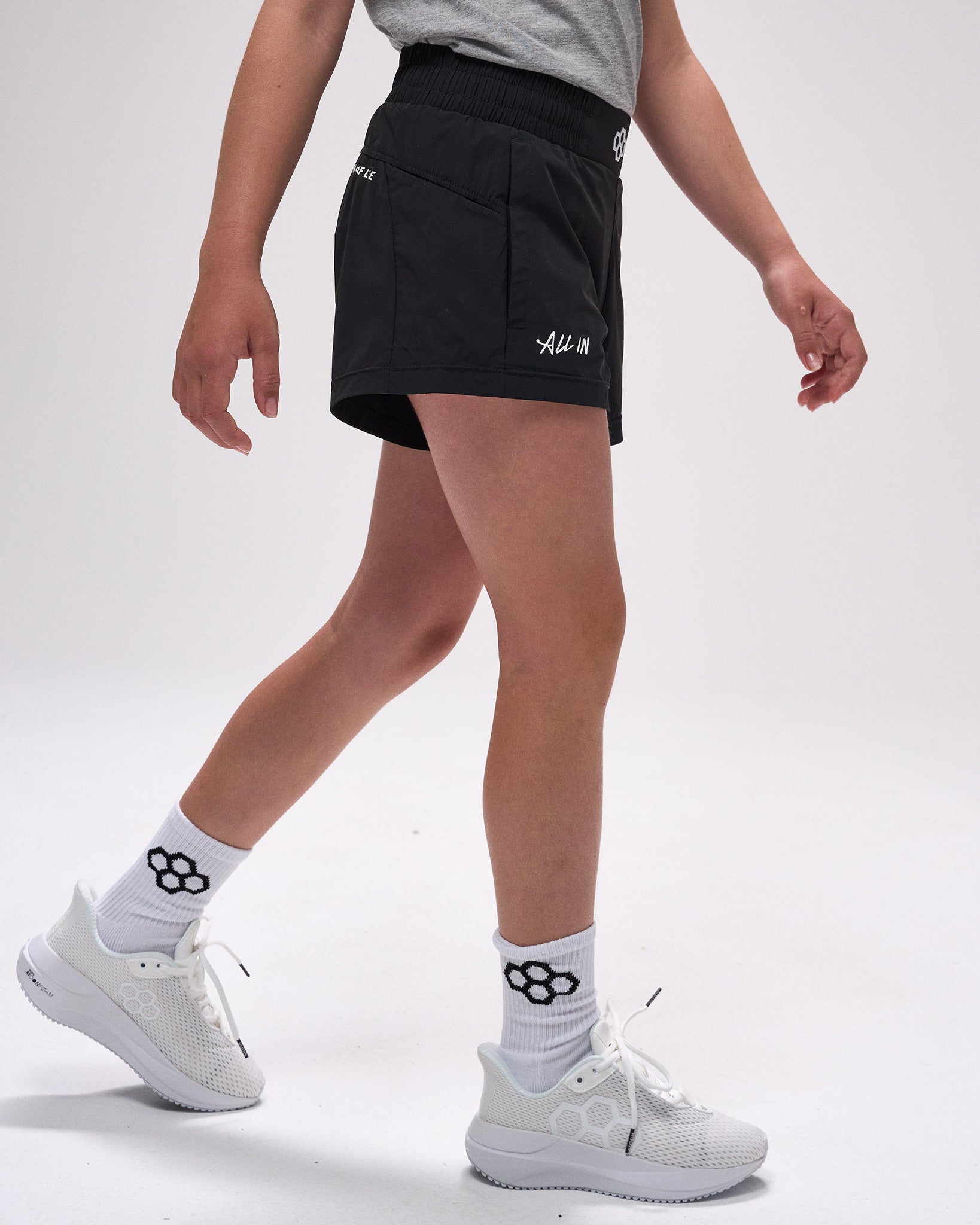 Person wearing black shorts with a logo, white sneakers, and socks on a light gray background