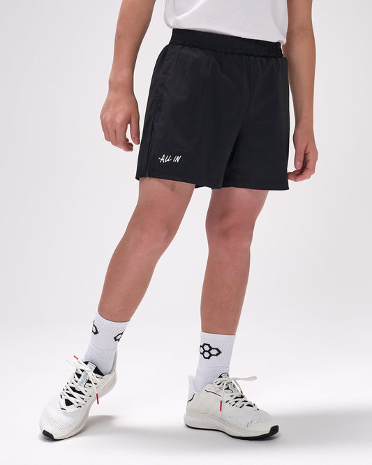 Person wearing black shorts with 'All In' text, white socks, and white sneakers on a light gray background