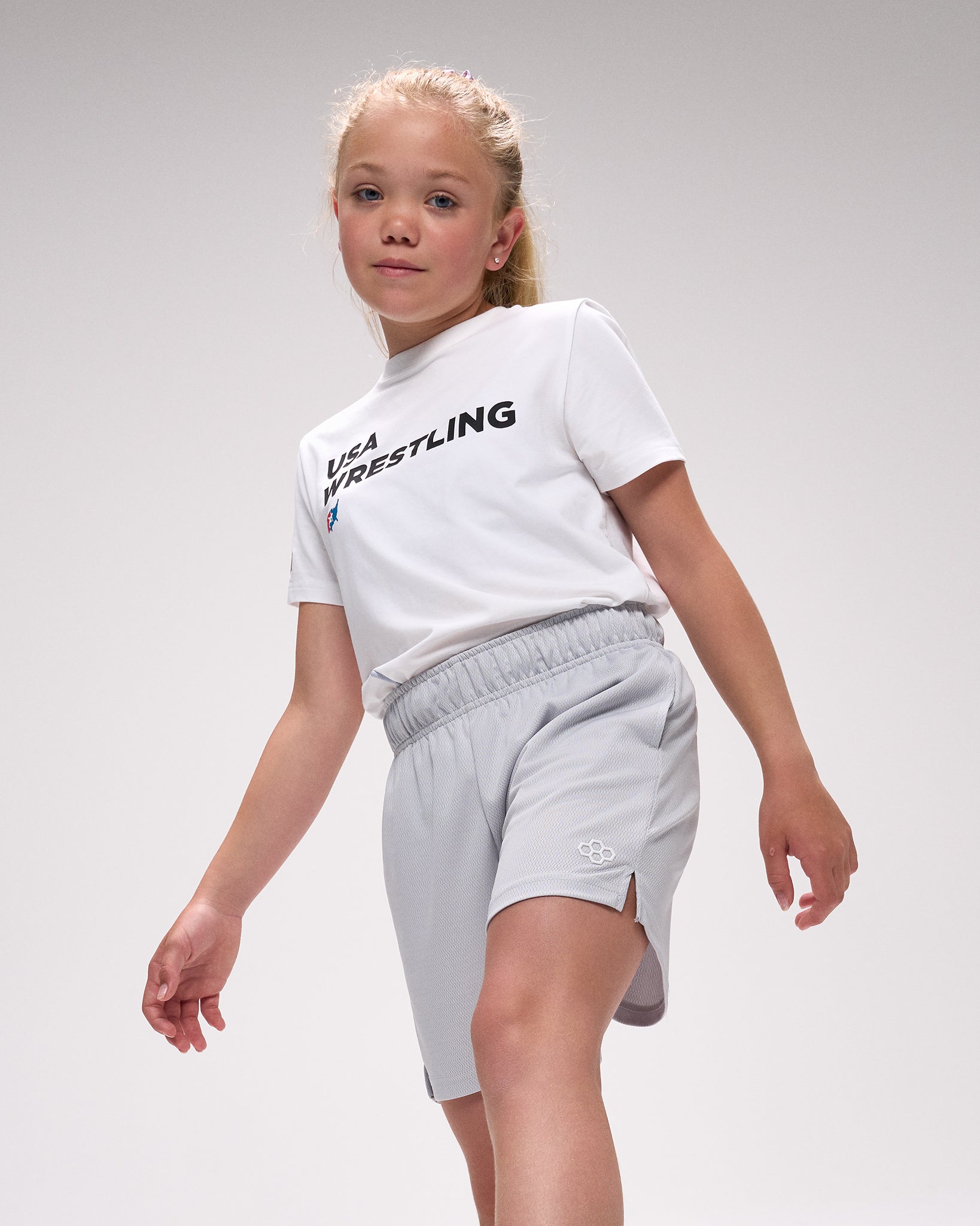 Child wearing a white 'USA Wrestling' t-shirt and gray shorts on a light gray background