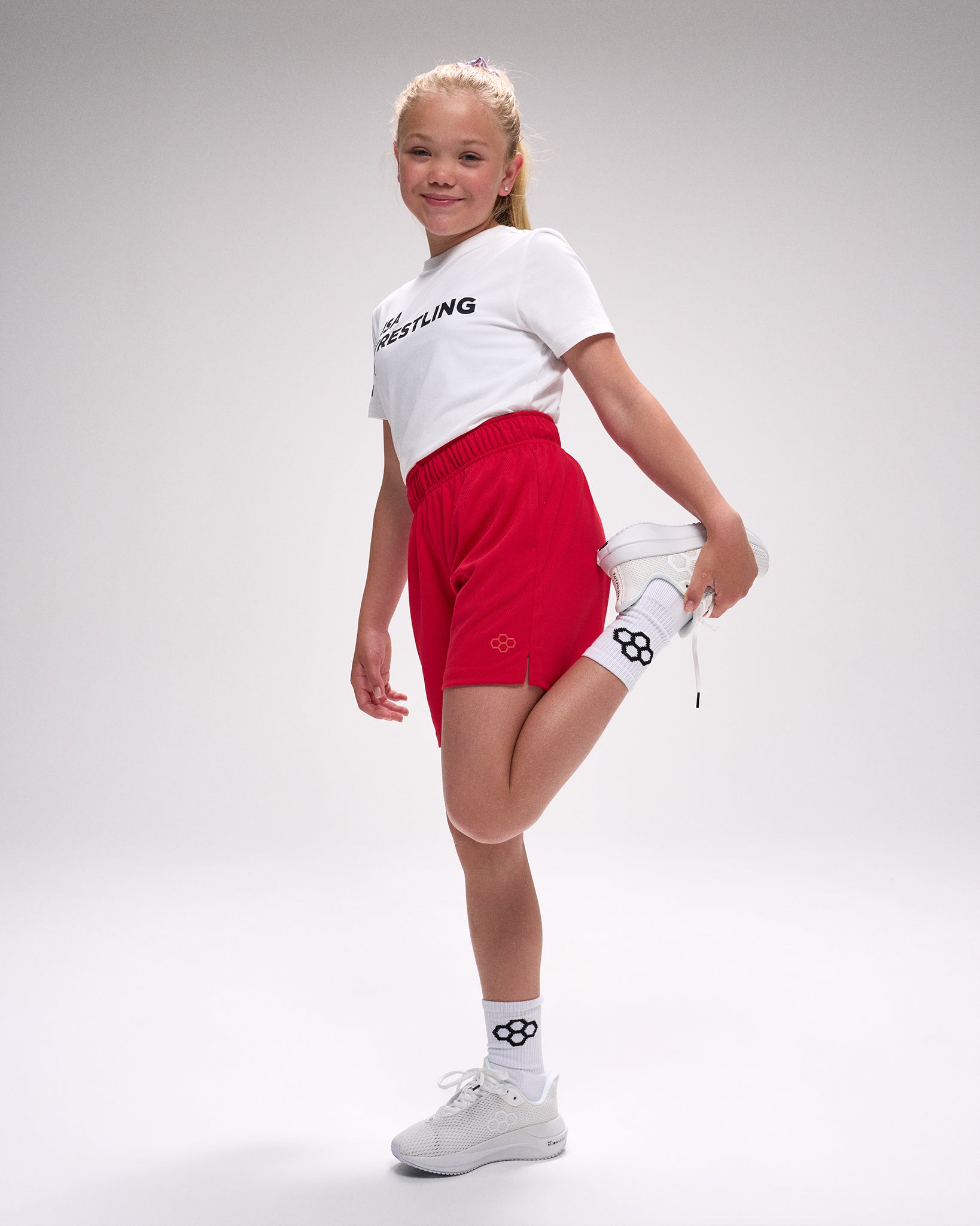 Young girl in white t-shirt with 'Wrestling' text, red shorts, and white sneakers on a gray background