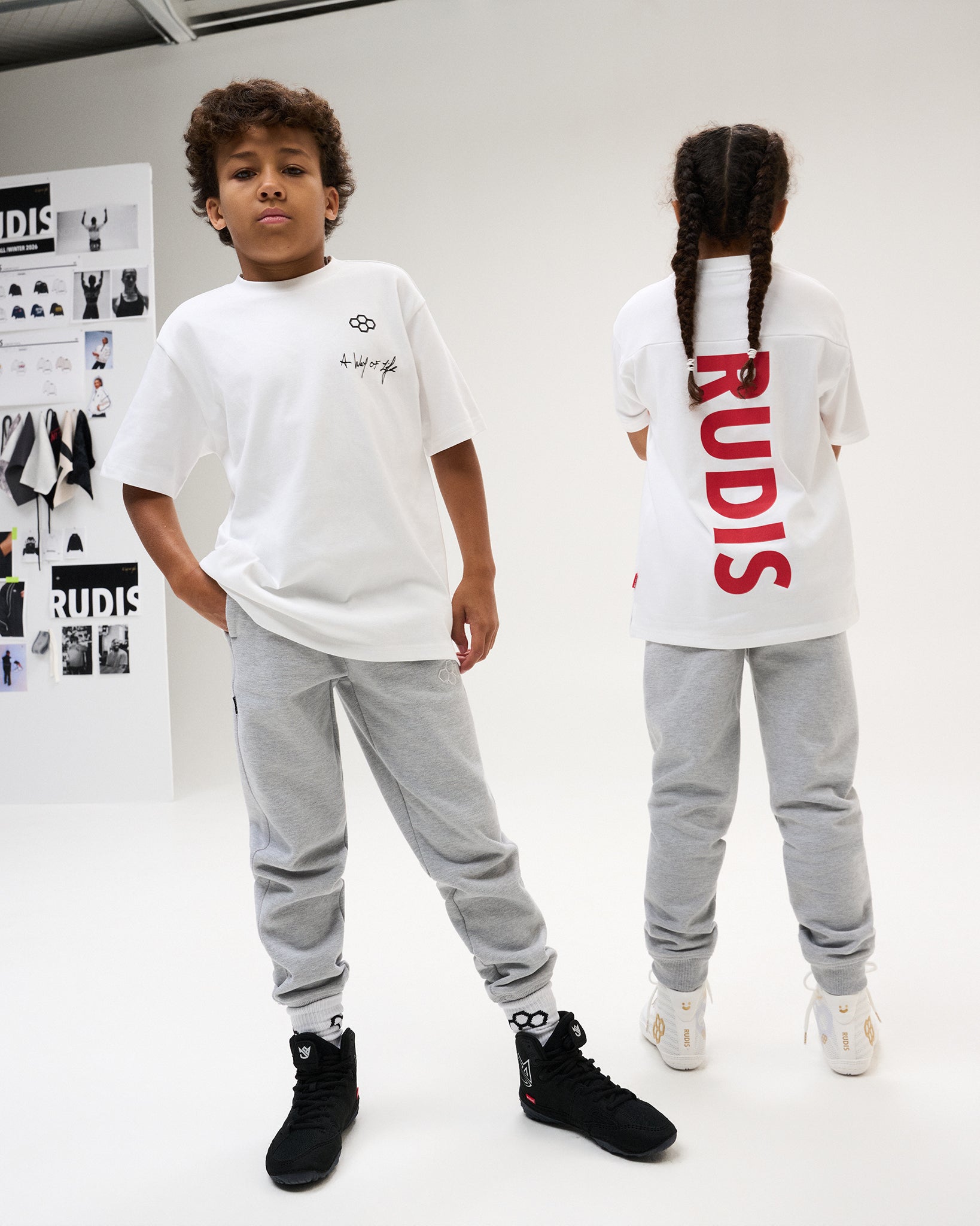 Youth athletes model front and back views of the white RUDIS Script Youth Drop Shoulder Tee with gray joggers in a studio setting.