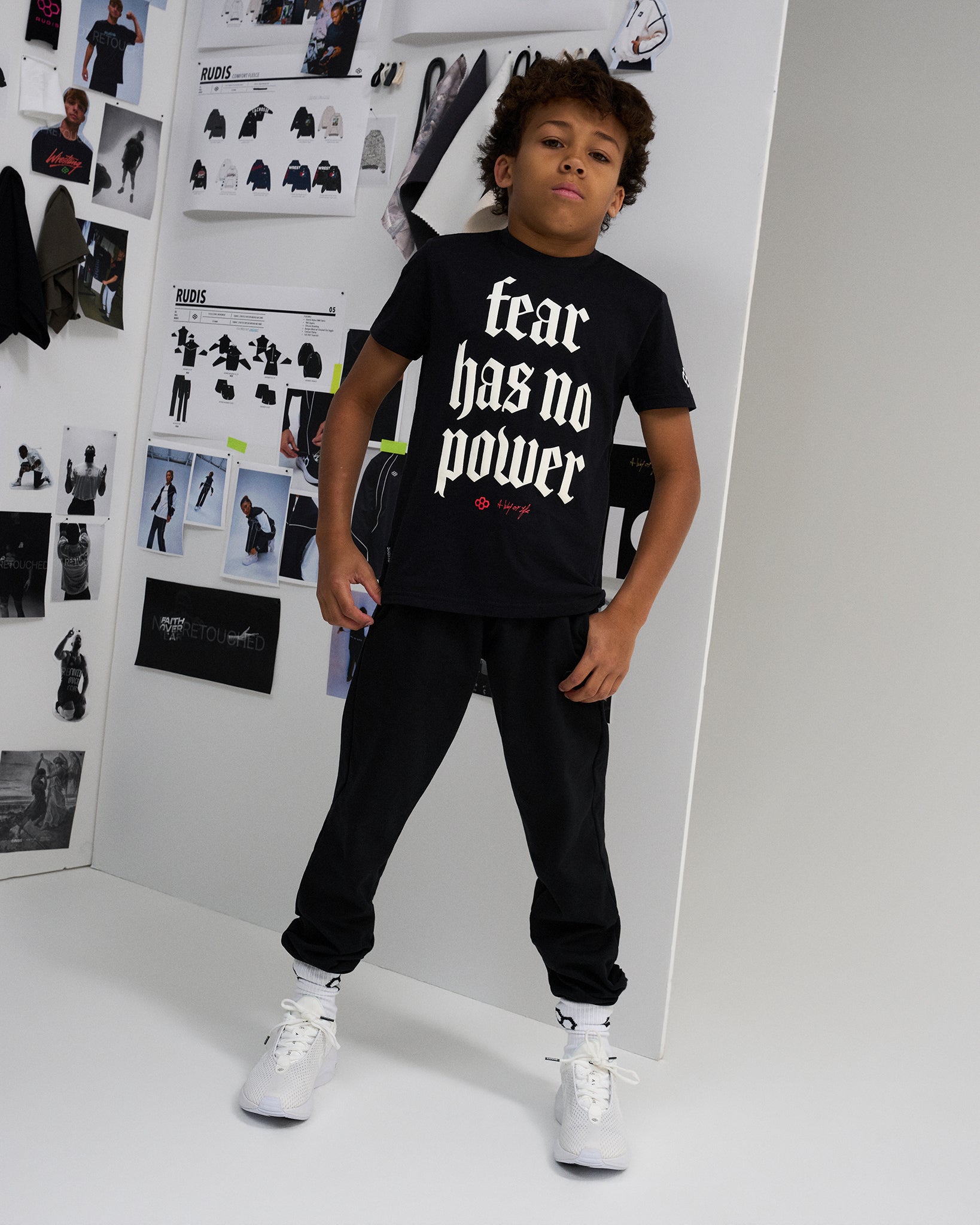 A child model wearing a black graphic t-shirt with the text 'fear has no power' in white gothic font, styled with black sweatpants and white sneakers, standing in a studio with fashion concept boards on the wall.