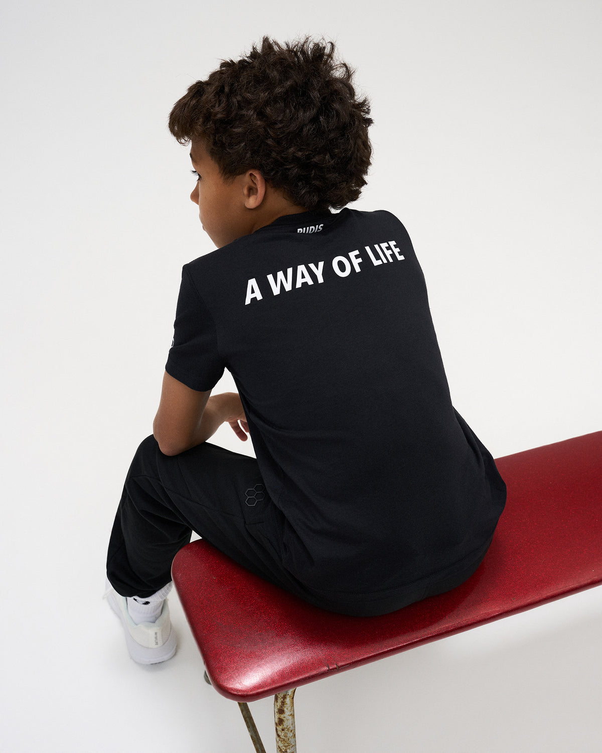A child wearing a black t-shirt with 'A WAY OF LIFE' printed in white on the back, sitting on a red bench against a white studio background.