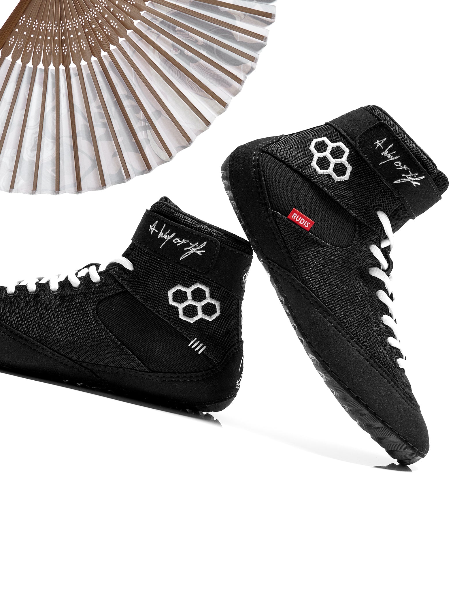 Black wrestling shoes with white laces and logo, next to a decorative fan on a white background
