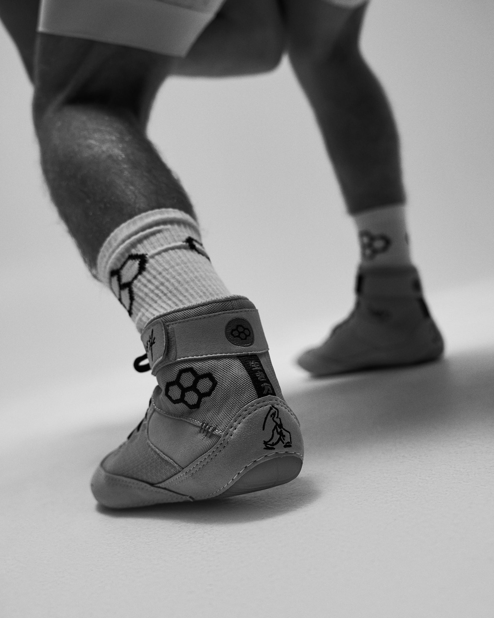 A close-up of a child's legs wearing a gray wrestling shoe, white socks with black geometric patterns, and gray leggings in a studio setting.
