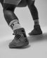 A close-up of a child's legs wearing a gray wrestling shoe, white socks with black geometric patterns, and gray leggings in a studio setting.