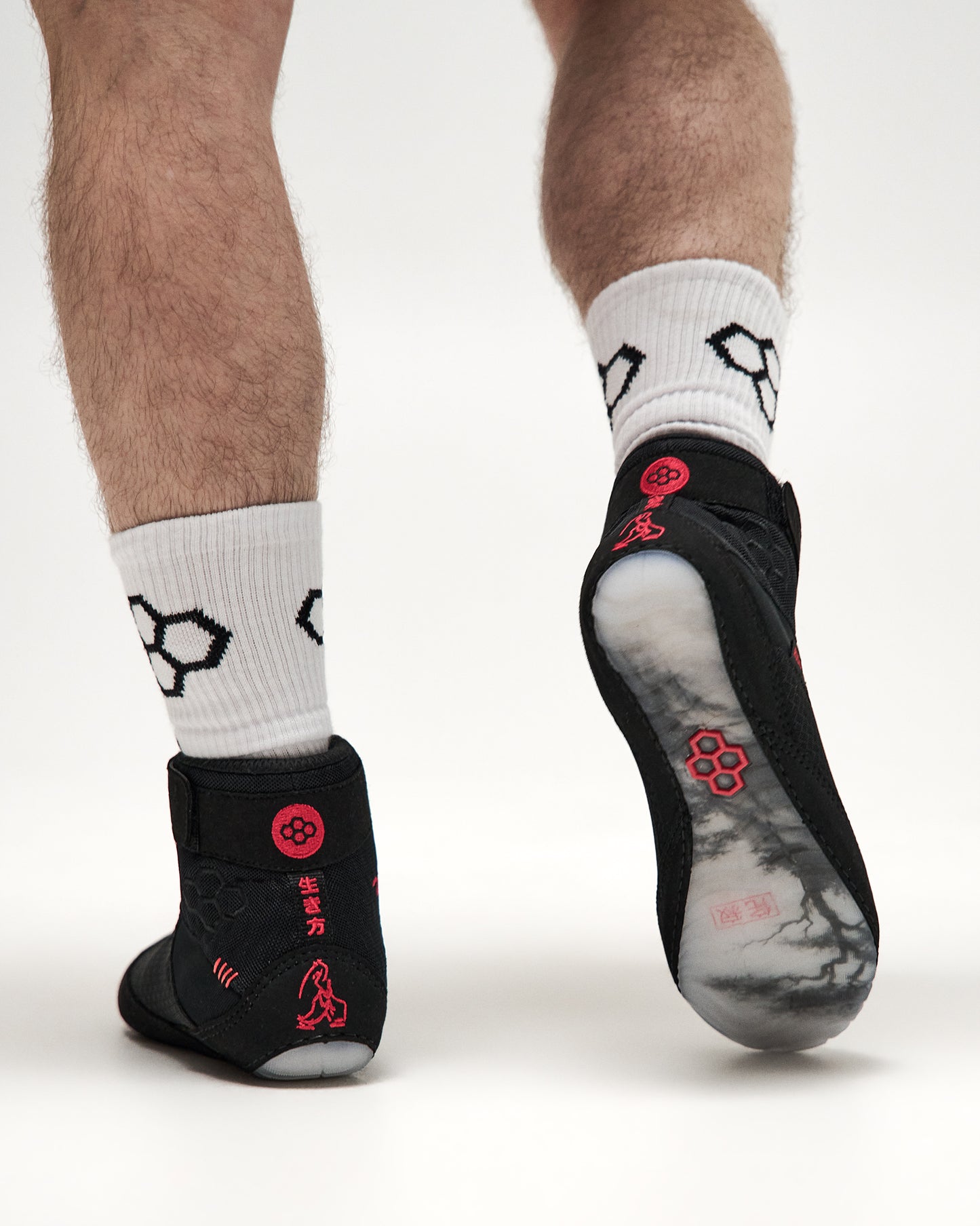 Black martial arts grip wrestling shoes worn on feet with white patterned crew socks, shown from the back and sole on a light gray studio background.