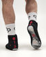 Black martial arts grip wrestling shoes worn on feet with white patterned crew socks, shown from the back and sole on a light gray studio background.
