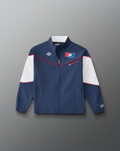 A navy blue zip-up track jacket with white shoulder panels and thin red piping details, featuring team-style graphics on the chest, photographed against a neutral gray background.