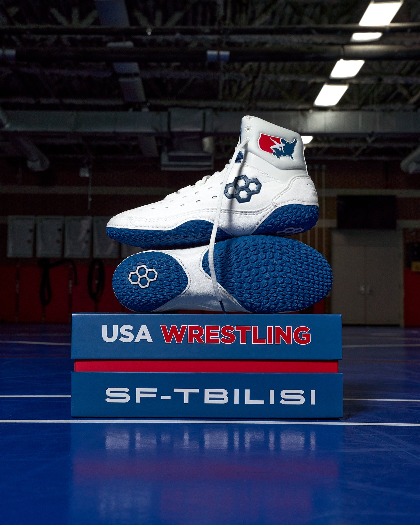 White and blue wrestling shoes on a blue platform with 'USA Wrestling' and 'SF-Tbilisi' text, in an indoor sports facility.