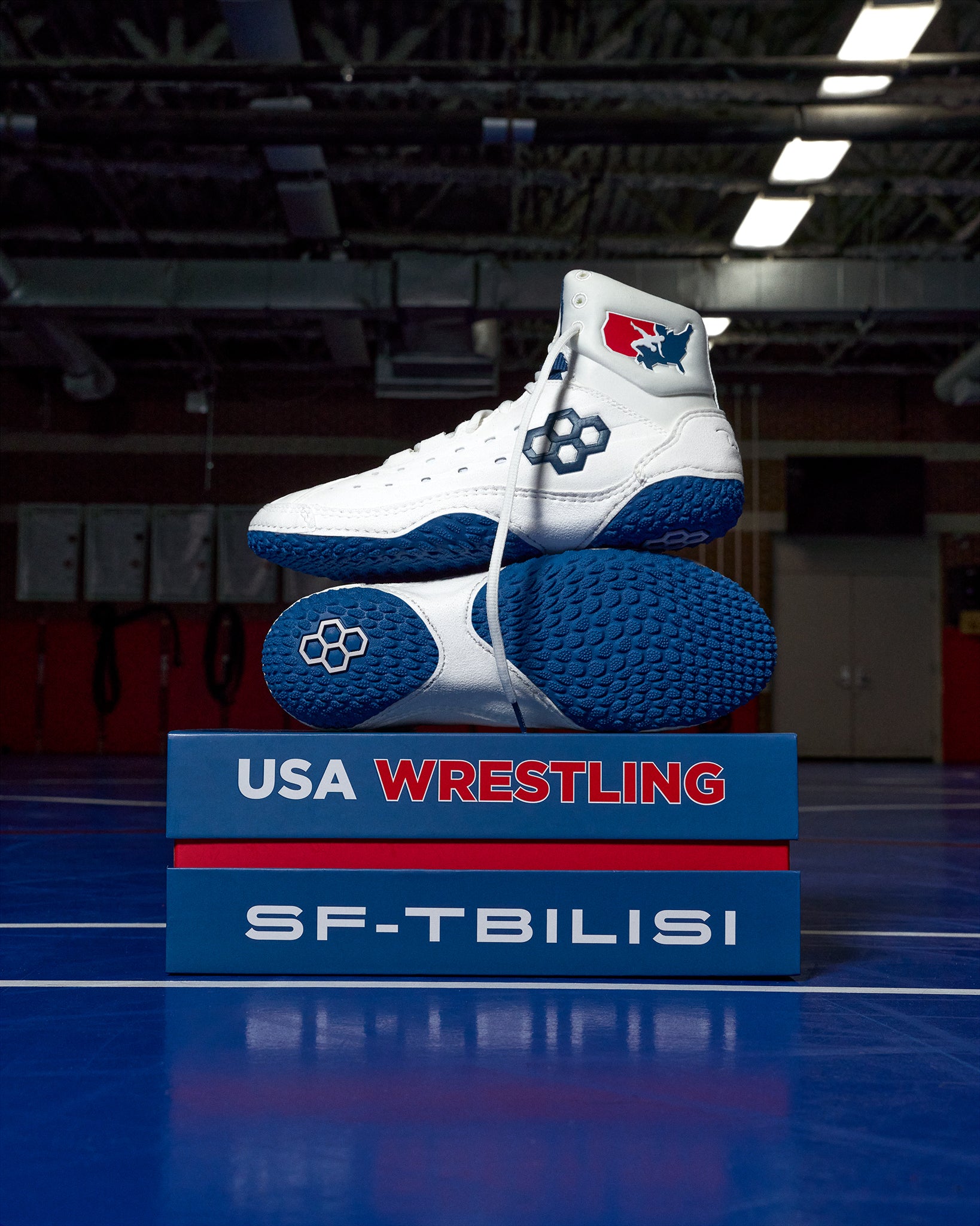 White and blue wrestling shoes on a blue platform with 'USA Wrestling' and 'SF-Tbilisi' text, in an indoor sports facility.