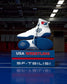 White and blue wrestling shoes on a blue platform with 'USA Wrestling' and 'SF-Tbilisi' text, in an indoor sports facility.