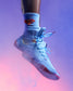 A light blue high-top wrestling shoe with matching blue laces and a red Superman logo on the side, worn with a light blue crew sock featuring a red and yellow Superman emblem. The shoe is shown mid-jump against a purple and pink smoky background.