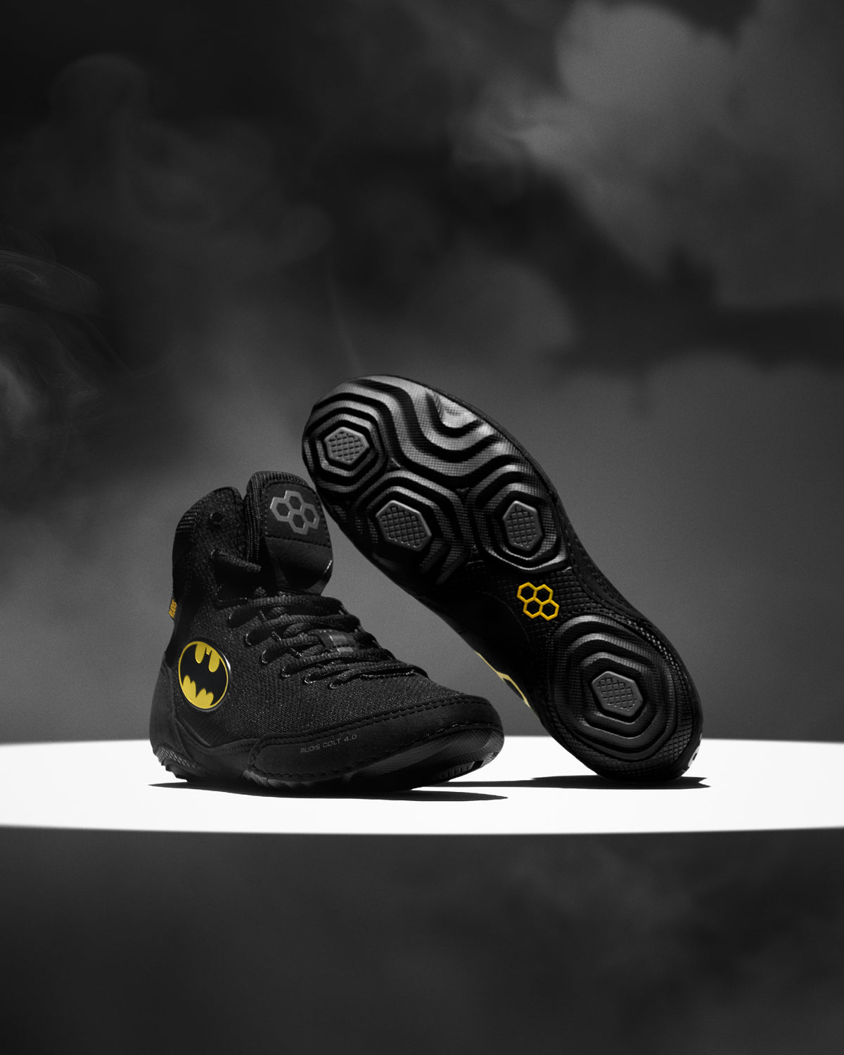 A pair of black high-top wrestling shoes with yellow Batman-themed accents, including a Batman logo on the side and a yellow circular emblem on the sole, displayed on a white platform against a dark smoky background.