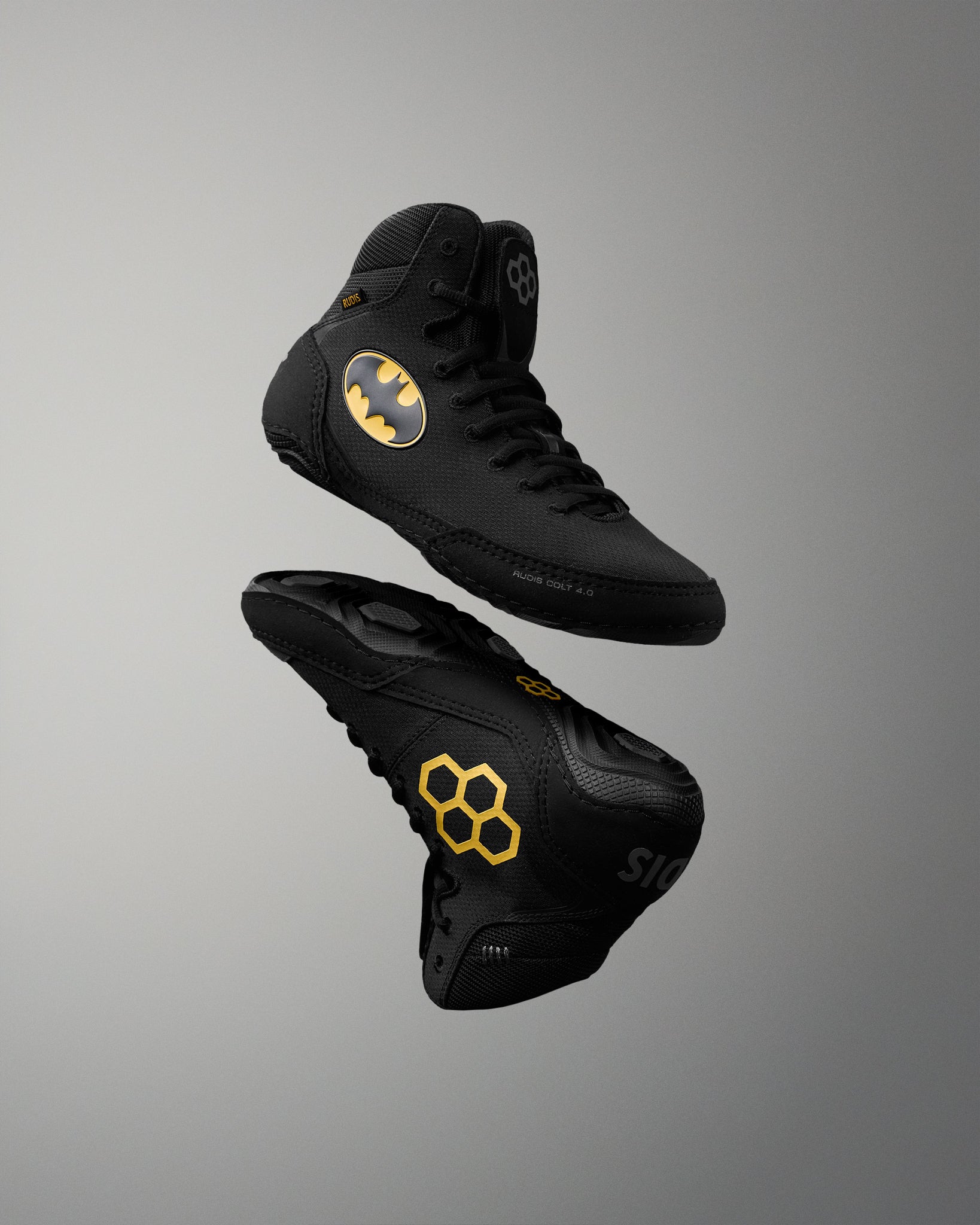 Two black high-top wrestling shoes floating against a gray gradient background. One shoe displays a yellow Batman logo on the side, and the other shows a yellow honeycomb pattern on the sole.