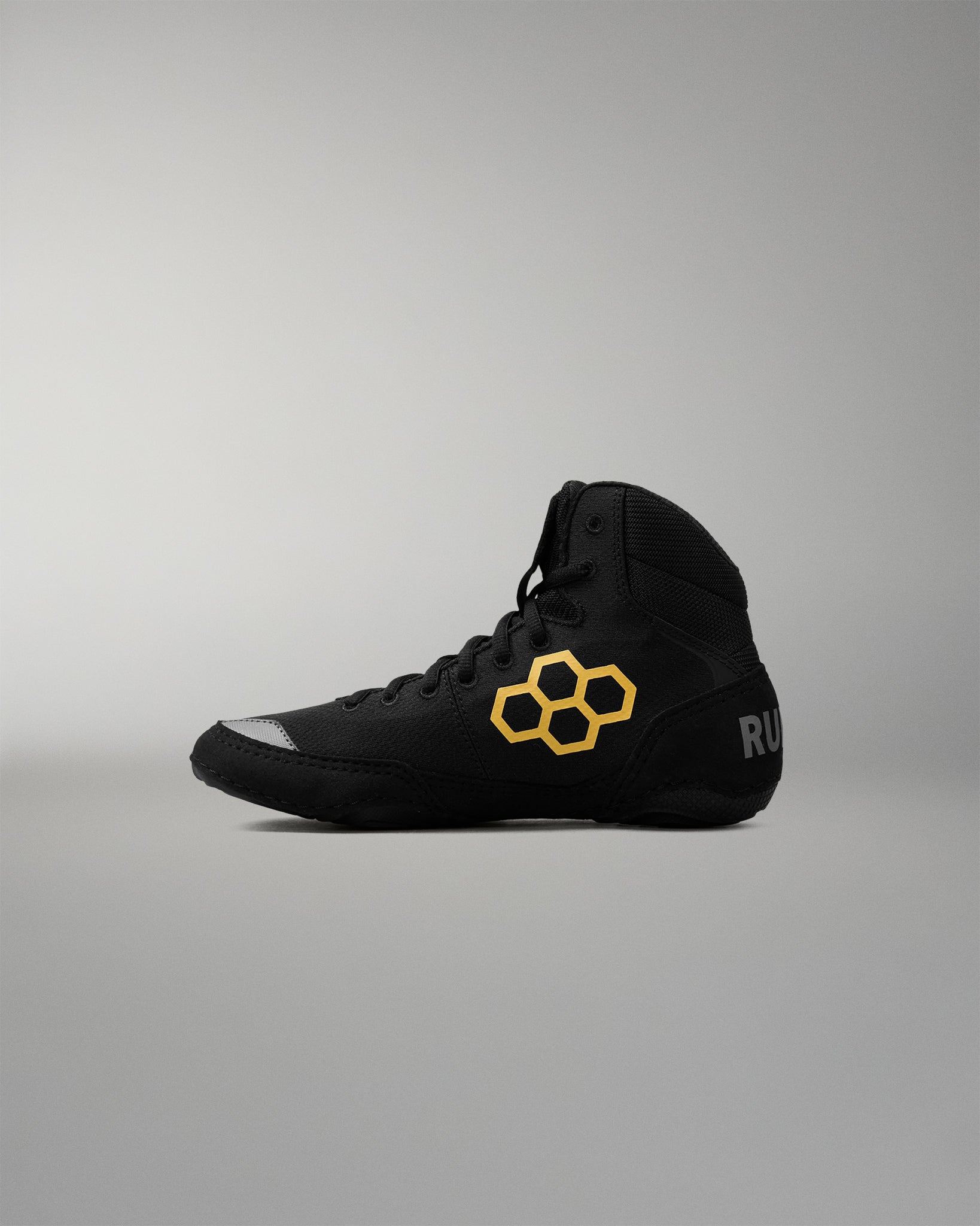 A black high-top wrestling shoe with a yellow geometric honeycomb logo on the side and a small gray logo on the heel, shown in profile against a neutral gray background.