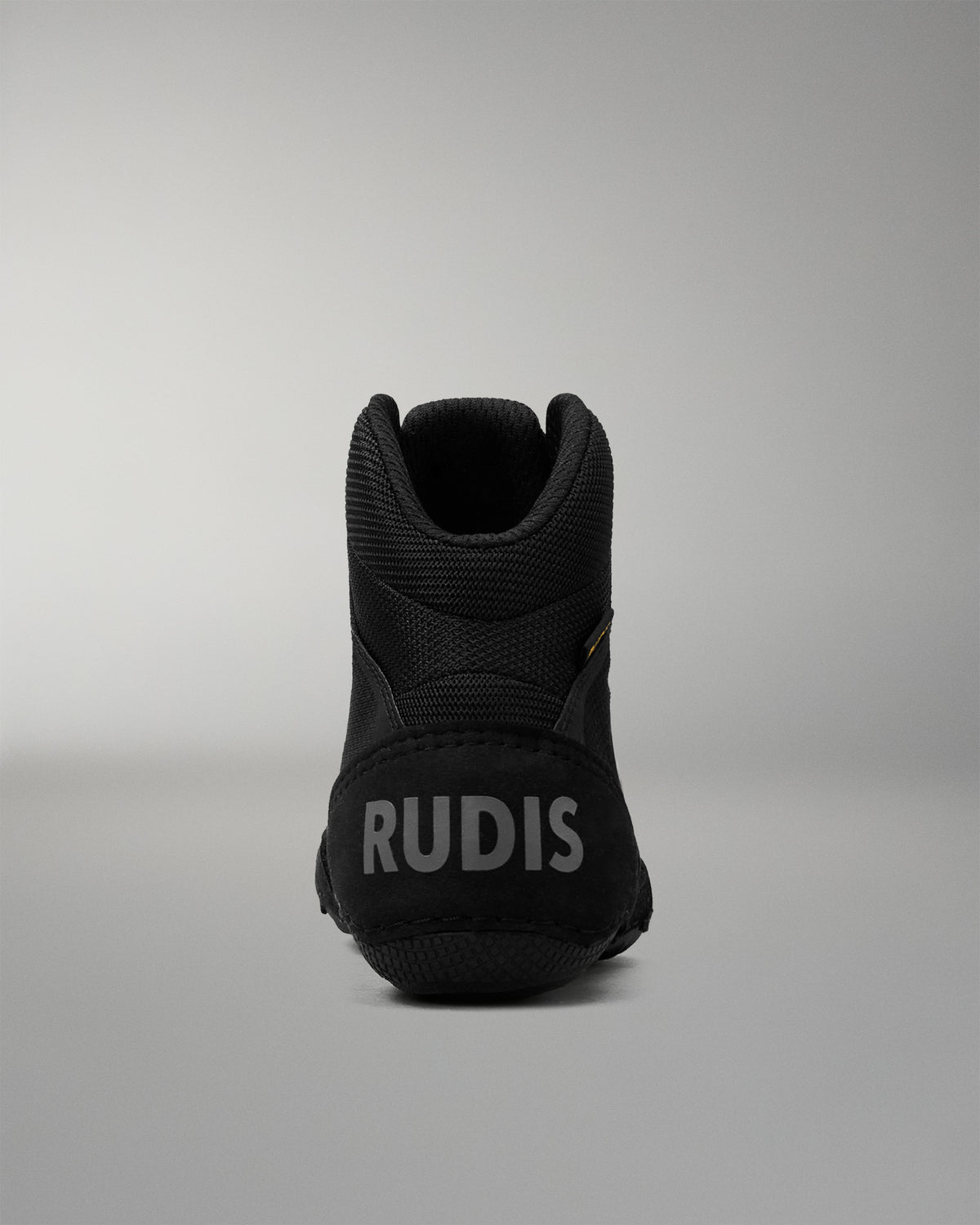 A black mid-top wrestling shoe shown from the rear, featuring the brand name 'RUDIS' in gray lettering on the heel, set against a neutral gray studio background.