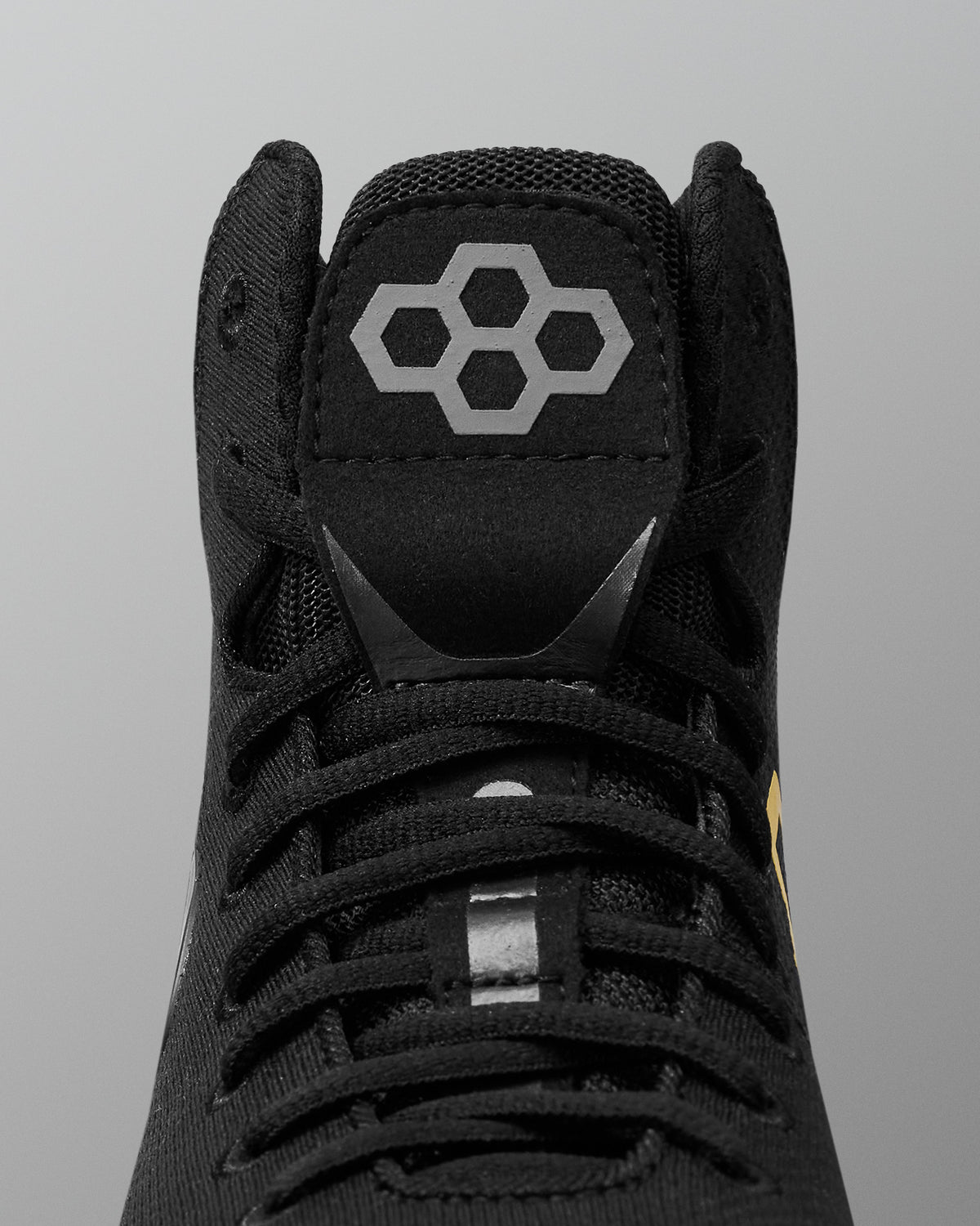 Close-up of a black high-top wrestling shoe showing the tongue with a gray geometric hexagon logo, black laces, and a small yellow accent near the side, set against a neutral gray background.
